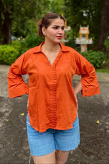 Woman wearing an orange shirt and blue shorts standing outdoors with greenery in the background