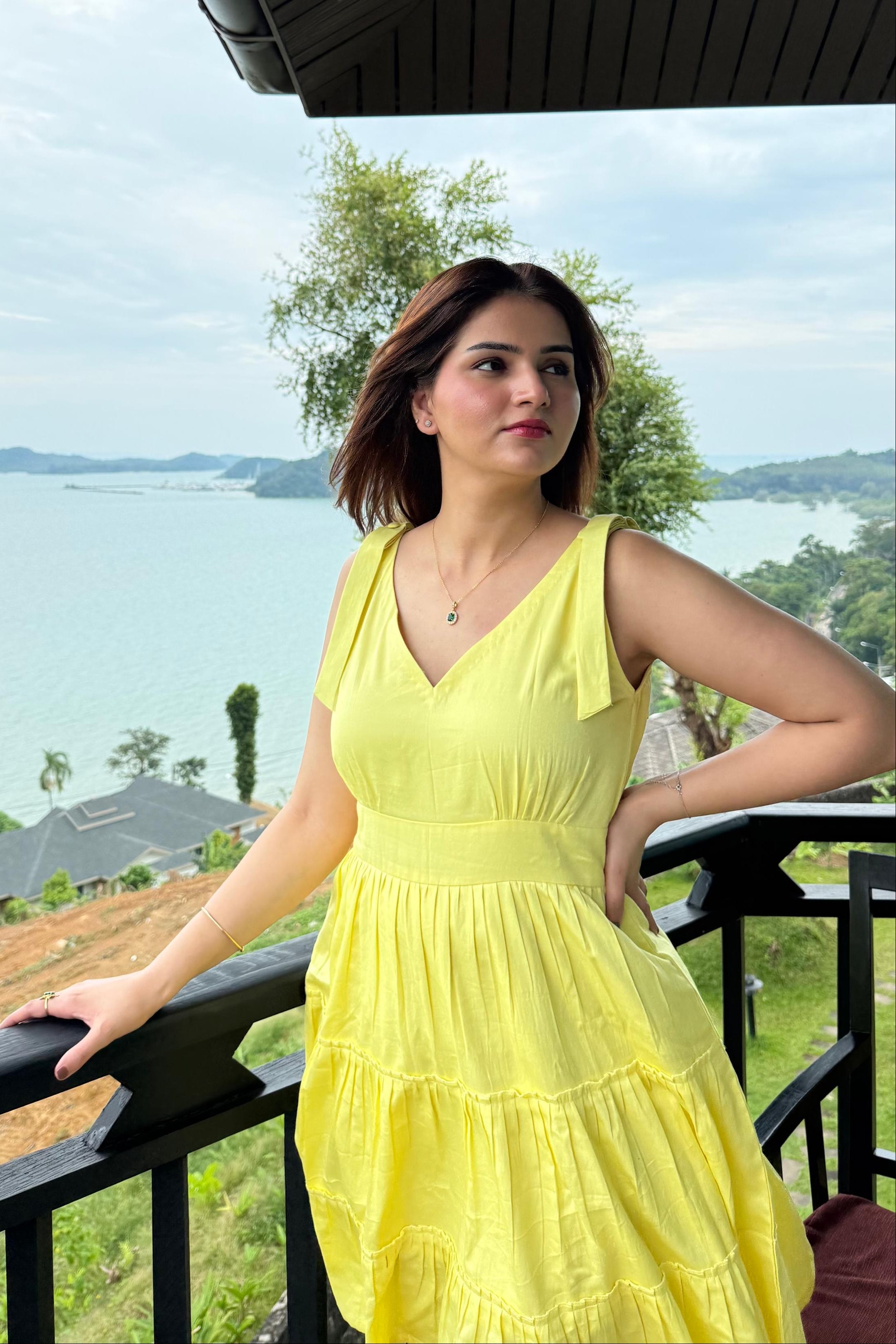 Woman in a yellow dress standing on a balcony with a scenic view of trees and water.
